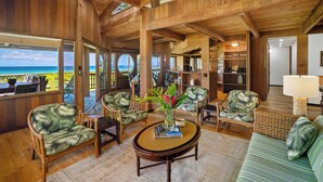 A spacious living area with wooden beams, a ceiling fan, and a view of the ocean. There are wicker chairs, a sofa, and a coffee table with books and a vase of flowers. - Wilikoki - a 6br/3ba beachfront home on Hanalei Bay (Hanalei)