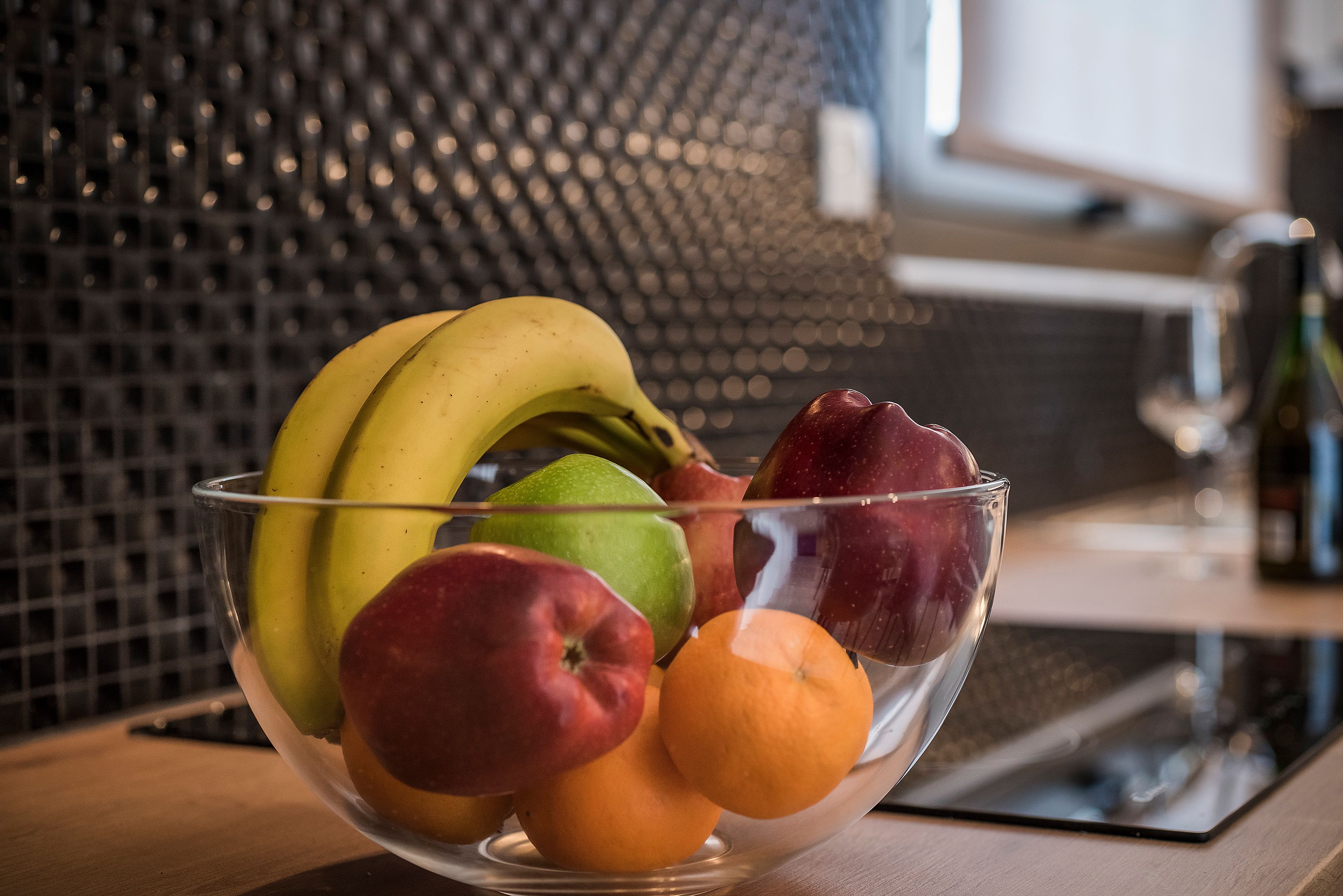A clear glass bowl containing various fruits including bananas, apples, and oranges, placed on a wooden surface with a tiled backsplash in the background.