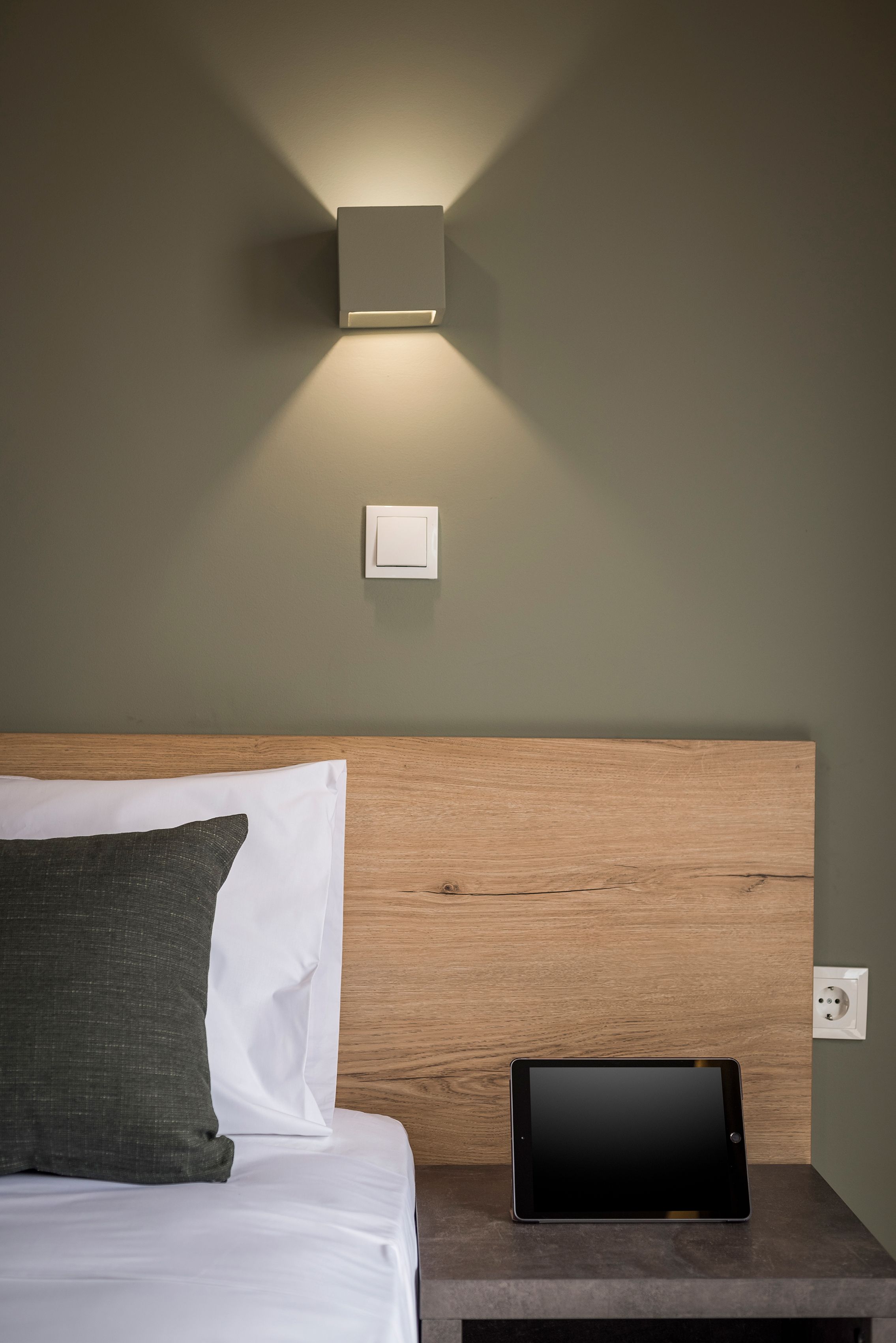 A neatly made bed with white and dark green pillows, a tablet on a bedside table, and a wall-mounted light fixture.