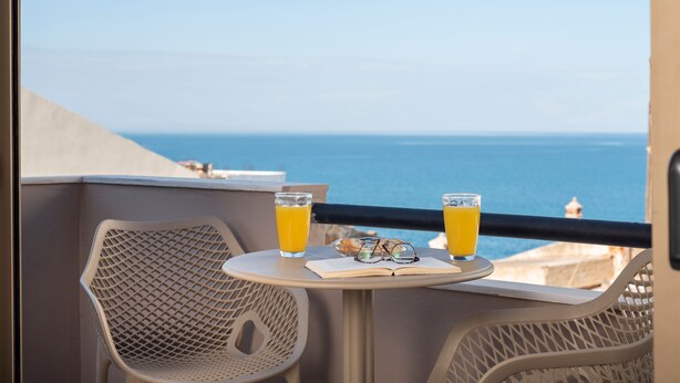 A balcony with a table, two glasses of orange juice, and a pair of glasses on a book. - Mirma Luxury Apartment (Chania)
