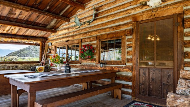 A rustic wooden cabin interior with a wooden table, benches, a window with a view of the mountains, and a door with a glass panel. - Double L Bar Lodge Guest Ranch (Cody)