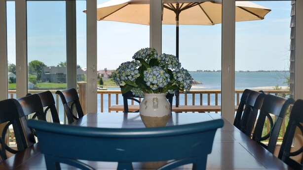 A dining area with a table and chairs, a vase with flowers, and a view of the water. - Luxurious Waterfront Retreat in Southampton: Breathtaking Views, Pool, and More! (Southampton)