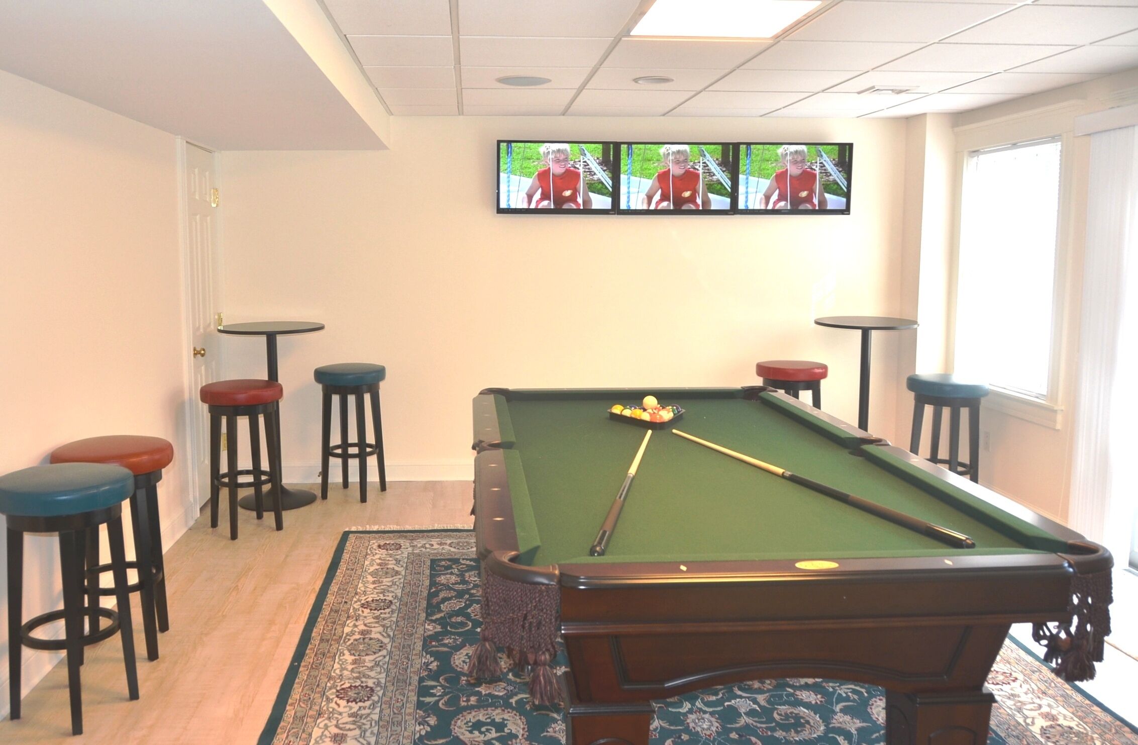 A pool table with stools, a television mounted on the wall, and a window with blinds.