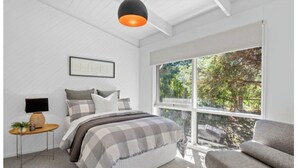 Ein Schlafzimmer mit Bett, Nachttisch mit Lampe, Fenster mit Blick auf Bäume und einem grauen Sofa.