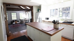 A kitchen with wooden cabinets, a countertop, and a window. There is a dining area with a table and chairs visible in the background. - Charming, Fully Restored, Greek Revival House, Farmland Setting (Buskirk)