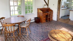 A dining area with a wooden table and chairs, a kitchen visible through an open door, and a brick floor. - Charming, Fully Restored, Greek Revival House, Farmland Setting (Buskirk)