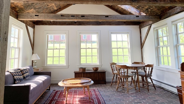 A rustic room with exposed wooden beams, a ceiling fan, and large windows offering a view of a red building and green lawn. - Charming, Fully Restored, Greek Revival House, Farmland Setting (Buskirk)