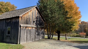A rustic wooden barn with a sloped roof, surrounded by a gravel driveway and a grassy area. - Charming, Fully Restored, Greek Revival House, Farmland Setting (Buskirk)