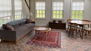 A cozy room with a sofa, a dining table with chairs, a wicker coffee table, and a patterned rug on a brick floor. - Charming, Fully Restored, Greek Revival House, Farmland Setting (Buskirk)
