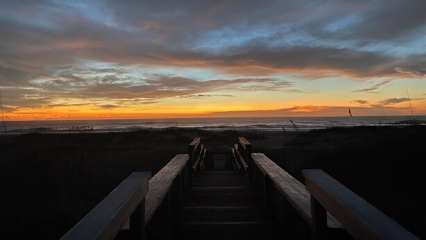 Ein hölzerner Steg führt zu einem Strand, im Hintergrund ist ein Sonnenuntergang zu sehen.