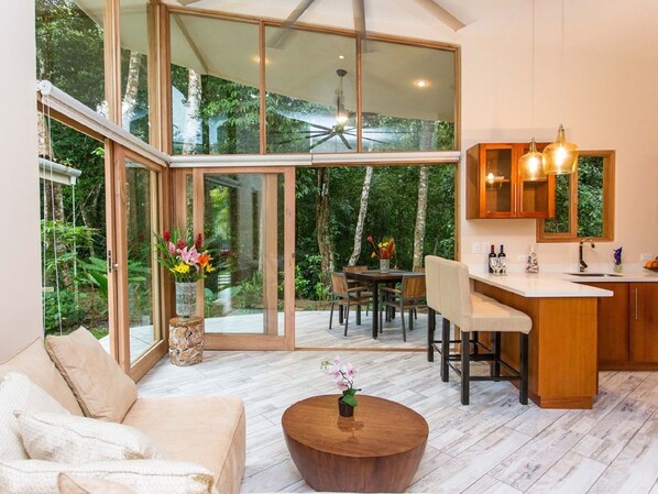A spacious living area with a large sliding glass door leading to a garden, a cozy seating area with a sofa and a coffee table, and a kitchen with wooden cabinets and pendant lighting. - 2 Bd Villa Mono Titi #3 at Rio Mono, Valle Pura Vida, Manuel Antonio (Quepos)