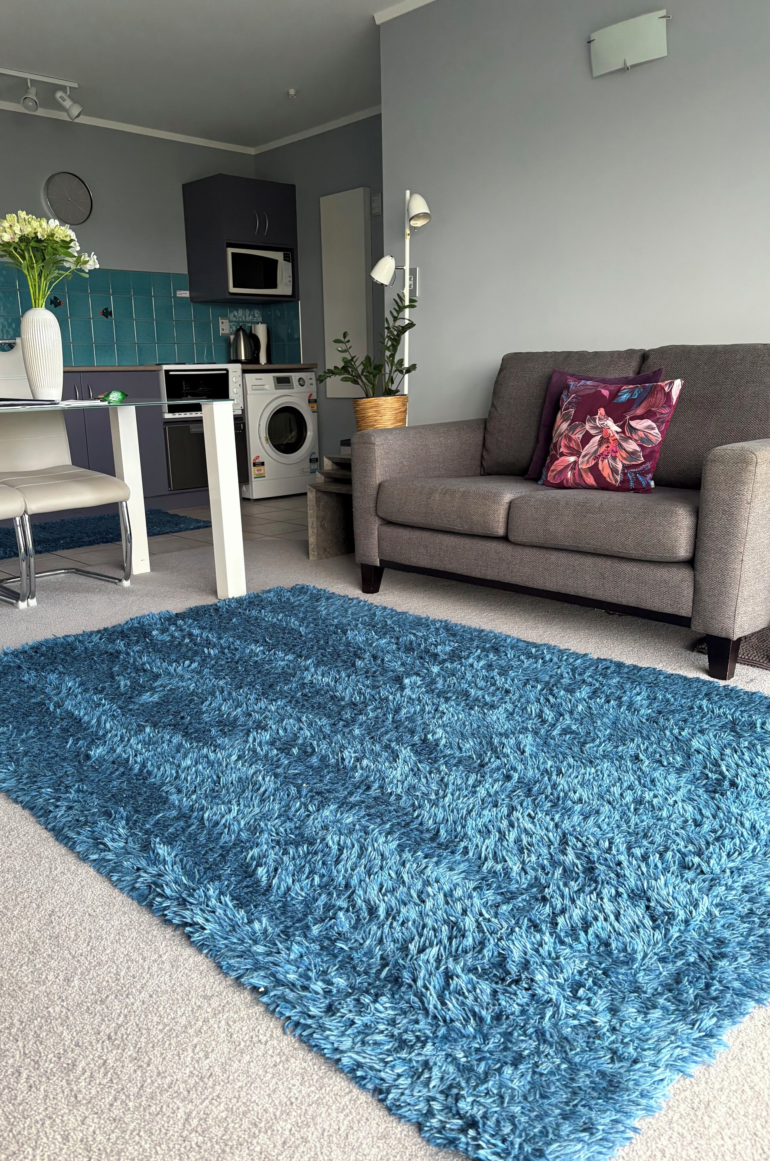 Una sala de estar con alfombra azul esponjosa, un sofá gris con un cojín floral, una mesa blanca y una cocina al fondo.