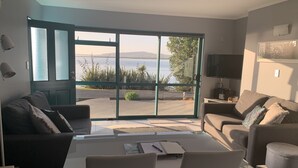 Een moderne woonkamer met een grote schuifpui die uitkomt op een balkon met uitzicht op een waterpartij en heuvels.