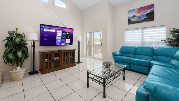 A living room with a flat-screen TV, a teal sofa, a glass coffee table, and a potted plant.