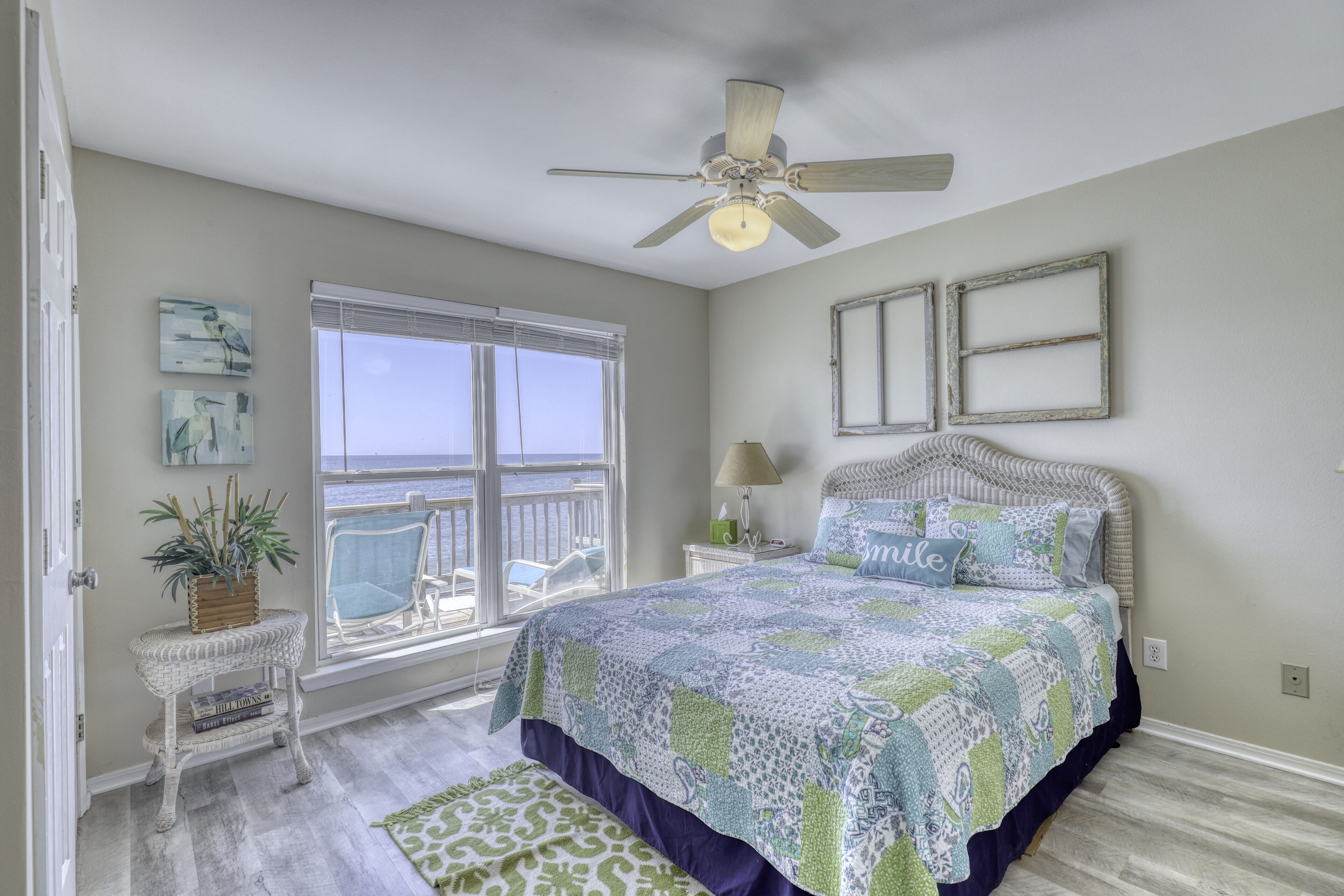 A bedroom with a large bed, a ceiling fan, a window with a view of the ocean, and a small side table with a plant.
