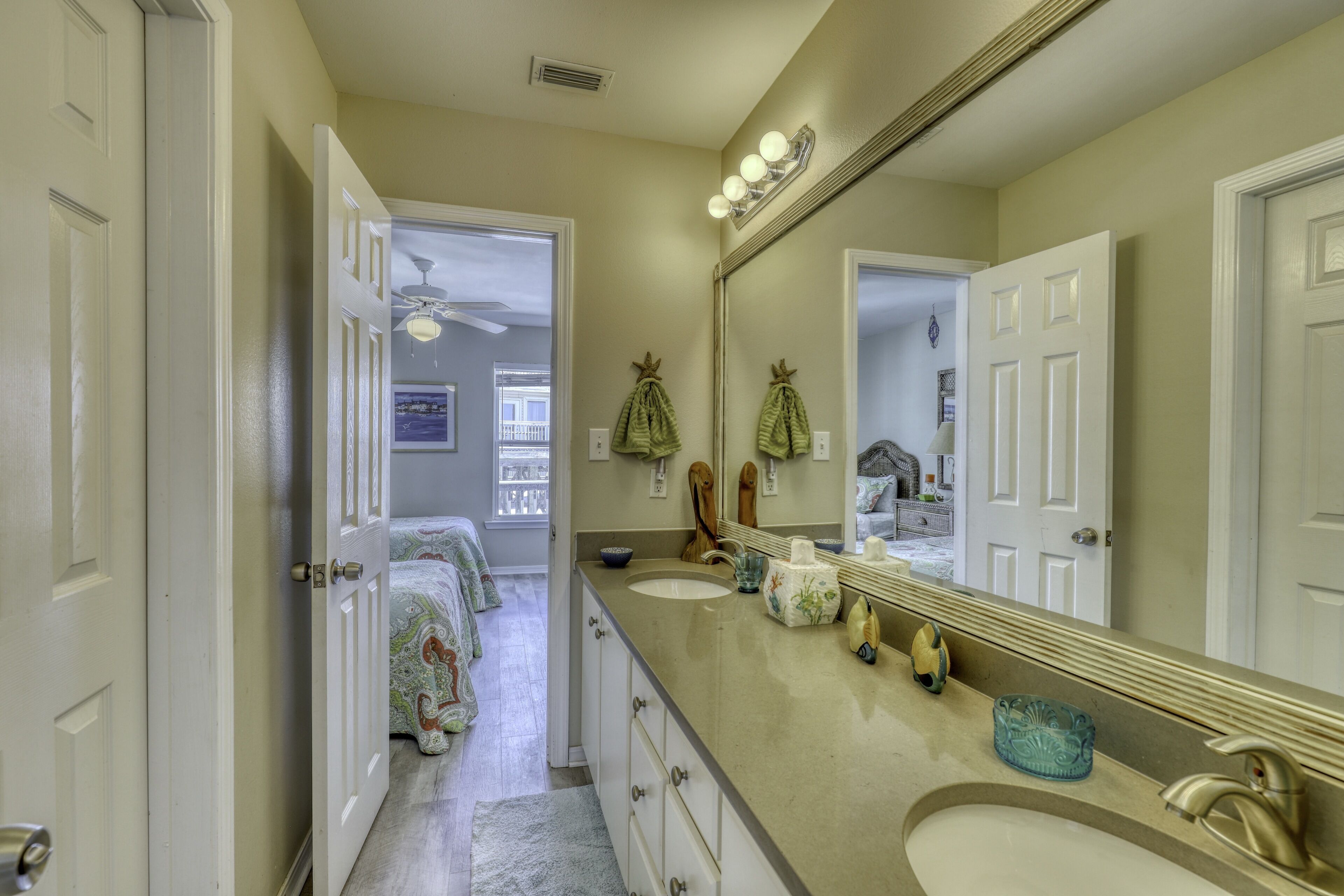 A bathroom with two sinks, a large mirror, and a door leading to a bedroom.