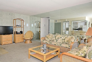 Sala de estar com televisão, móveis de vime, sofá com estofamento floral e cozinha visível por uma porta aberta.