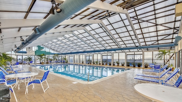 Area piscina interna con poltrone a sdraio e tavoli azzurri, pavimento piastrellato e soffitto in vetro.