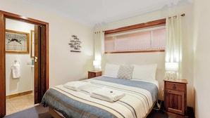 Un dormitorio con cama, mesitas de noche, una ventana con cortinas, un adorno en la pared y una puerta que da al baño.