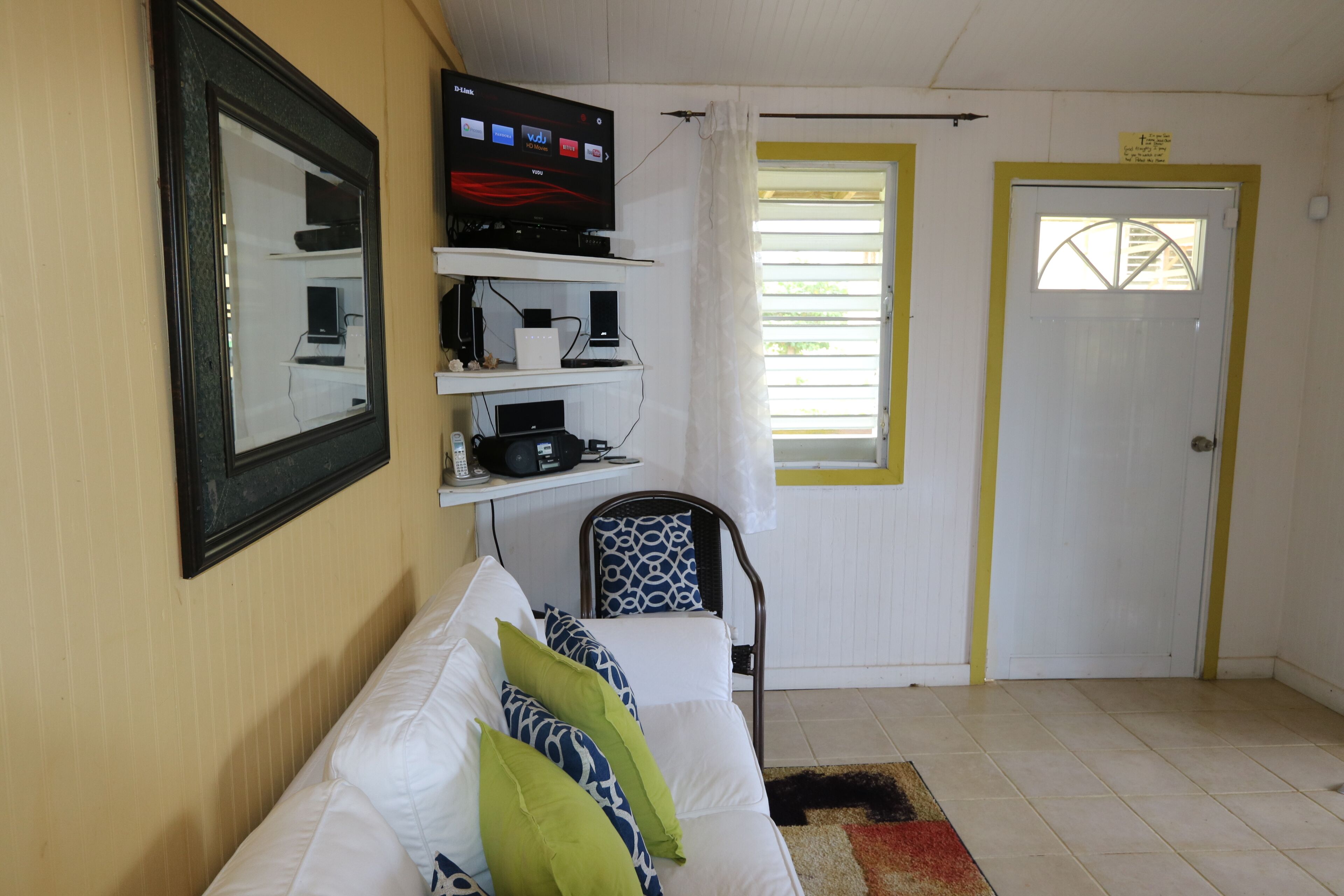 A hotel room with a white sofa, a television mounted on the wall, a door with a window, and a small shelf with electronic devices.