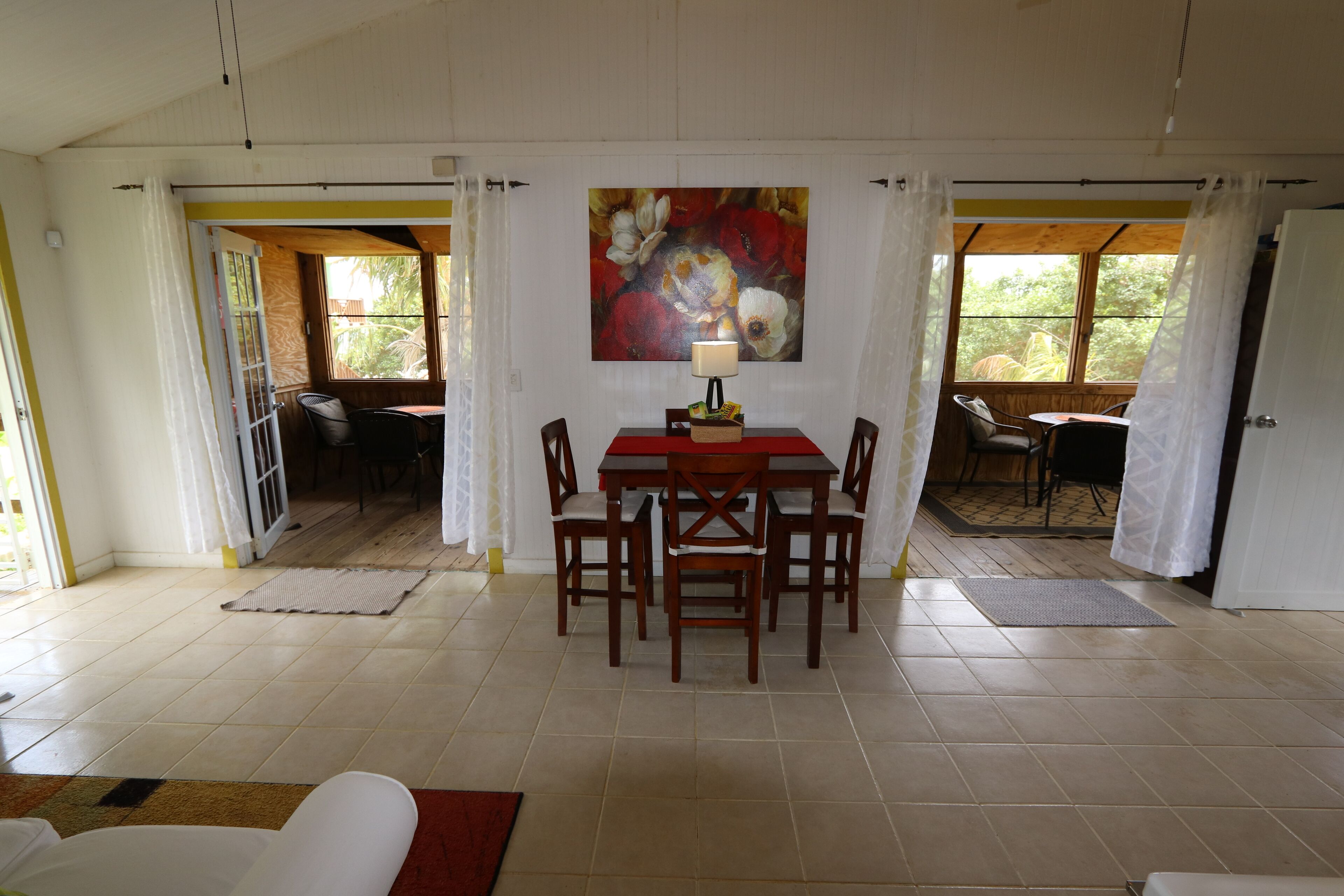 A dining area with a red table and chairs, a painting on the wall, and a view through the open door.