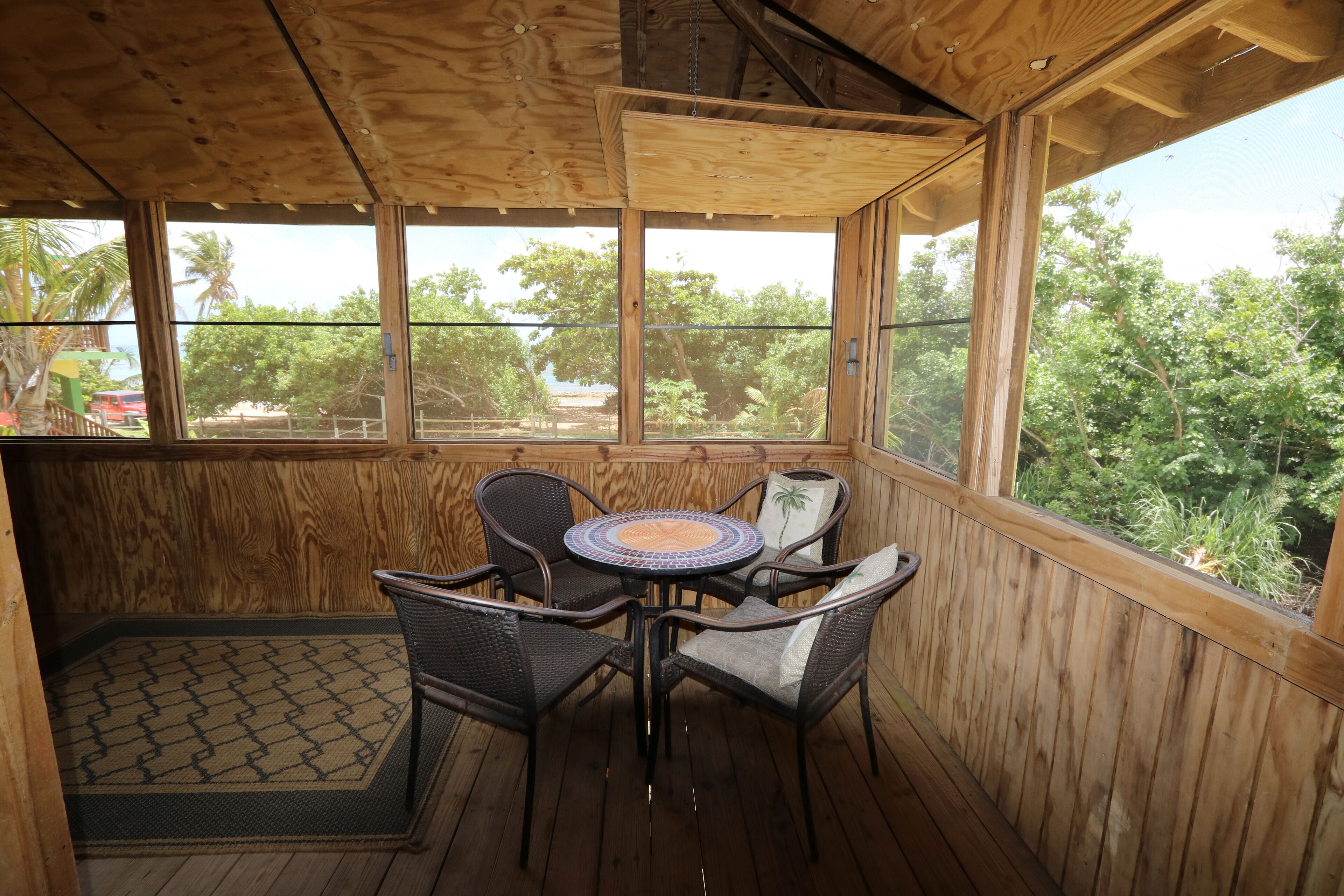 A wooden deck with a round table and four chairs, large windows, and a view of trees and a red car outside.