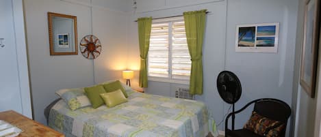 Een slaapkamer met een bed, een stoel, een bureau, een ventilator, een lamp en een raam met gordijnen.