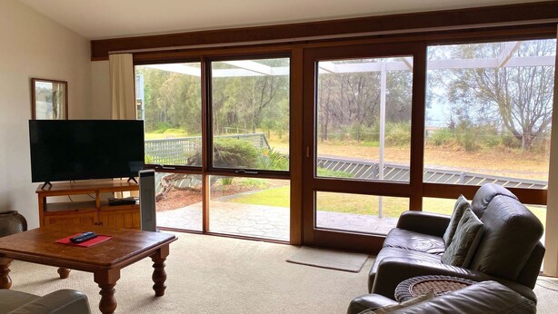 Un salon avec un téléviseur posé sur un meuble en bois, un canapé en cuir et une grande porte-fenêtre coulissante donnant sur une terrasse avec vue sur les arbres et une plateforme en bois.