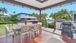 Ein überdachter Außenbereich mit Tisch, Stühlen, einem Grill und Blick auf das Meer und Palmen.