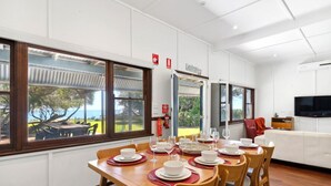 A dining area with a table set for a meal, large windows with a view of the outdoors, and a white sofa in the background. - Beach Cottage in the Heart of Mandurah (Mandurah)