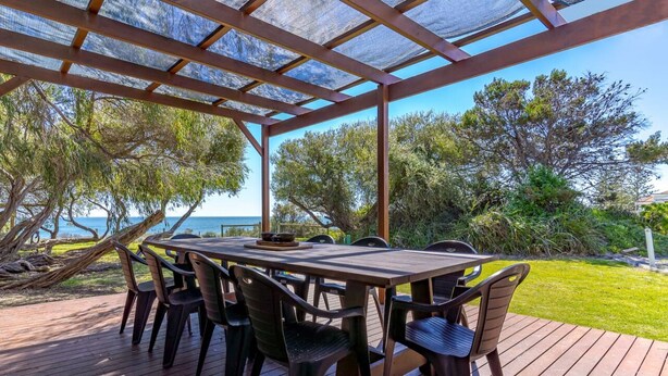 An outdoor patio with a wooden table and chairs, surrounded by trees and a view of the sea. - Beach Cottage in the Heart of Mandurah (Mandurah)