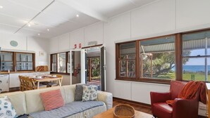 A spacious living area with a sofa, a red armchair, and a dining table. A large window offers a view of the outdoors. - Beach Cottage in the Heart of Mandurah (Mandurah)