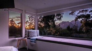 Ein modernes Zimmer mit großem Fenster und Blick auf eine gebirgige Landschaft. Ein Bett mit weißem Kopfteil, ein kleiner runder Tisch und eine Bank mit Kissen sind vorhanden.