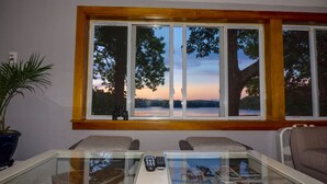 Habitación con un amplio ventanal con vistas a un lago al atardecer, una mesa de vidrio con controles remotos y una planta en maceta.