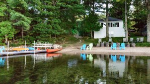 Una cabaña junto al lago con muelle, rodeada de árboles y una pequeña terraza con sillas.