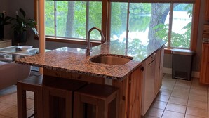 Una cocina con encimera de granito, un fregadero y una isla de madera. Hay un reloj en la pared y una ventana con vistas a árboles.