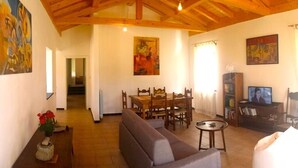 A spacious living area with a high wooden ceiling, a dining table with chairs, a sofa, and a television.