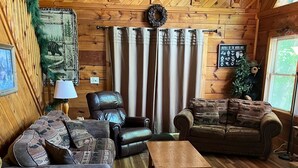 Ein gemütliches Blockhaus-Interieur mit Holzwänden und -böden, ausgestattet mit einem Ledersofa, einem Holzschaukelstuhl, einem Holztisch und einem gestreiften Sofa.
