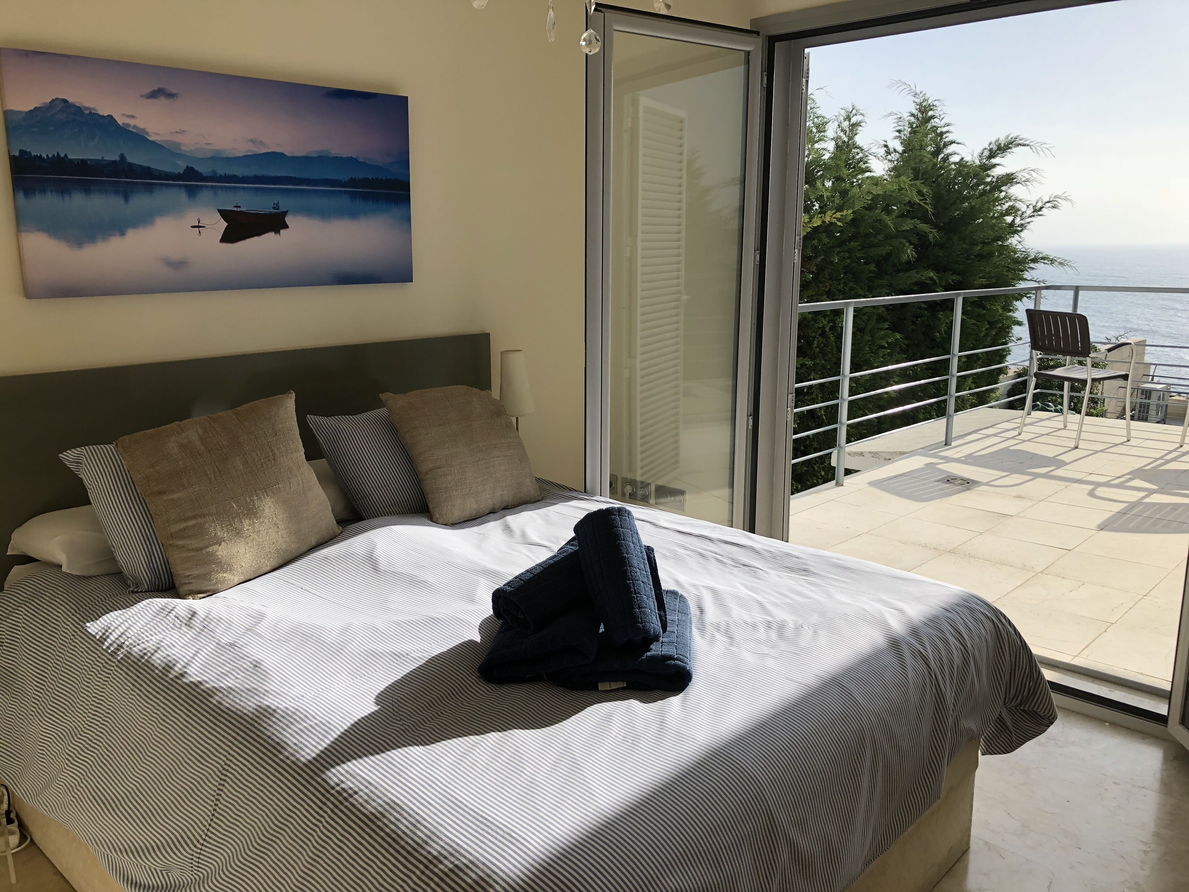 A bedroom with a bed, pillows, a painting, and a balcony with a view of the sea.