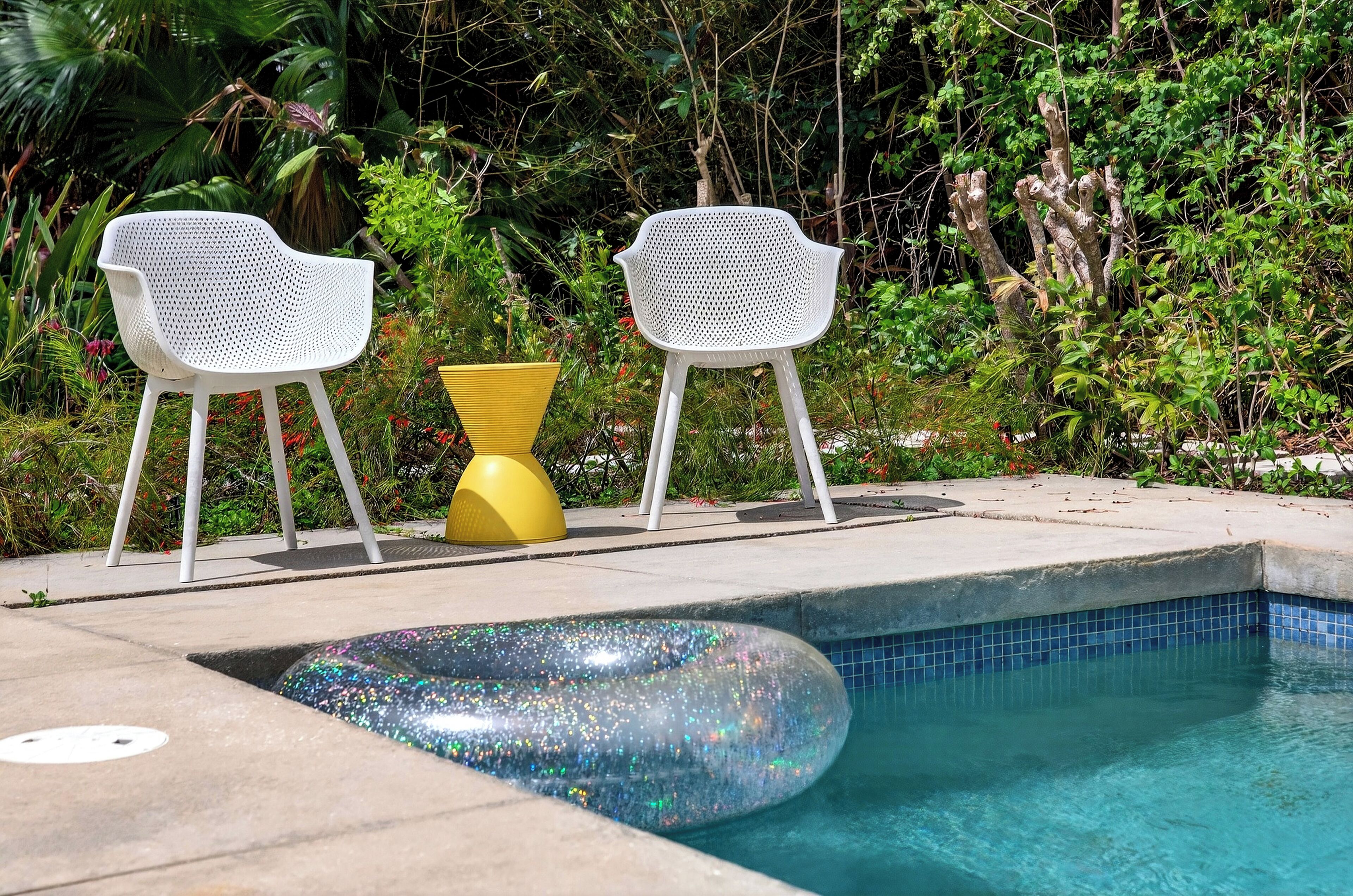 A pool area with two white chairs and a yellow table, surrounded by lush greenery.