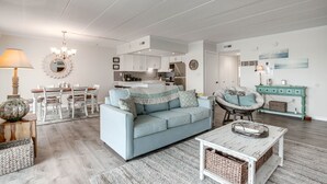 Un salon spacieux avec un canapé bleu clair, un coin repas avec des chaises blanches et une cuisine à l’arrière-plan.