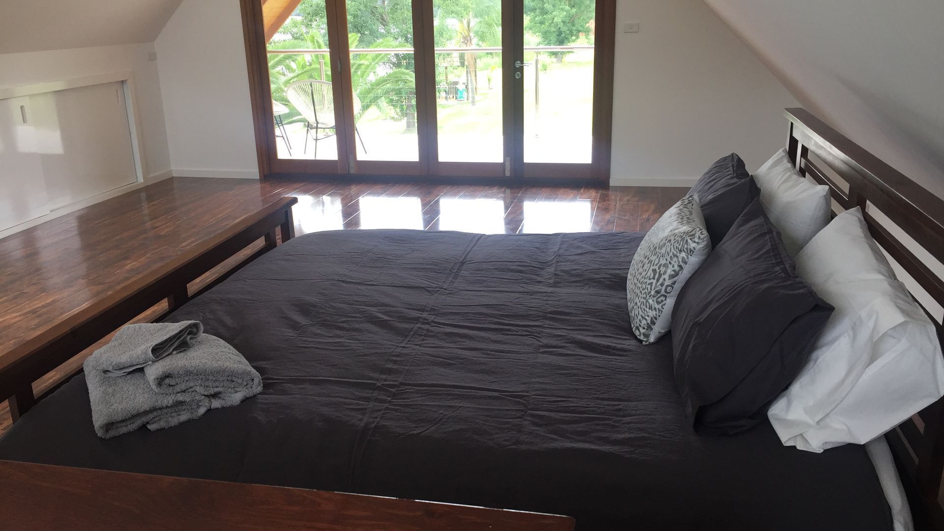 Un dormitorio con una cama grande, cabecero de madera y vistas a un espacio exterior con sillas y vegetación a través de una puerta corrediza de vidrio.