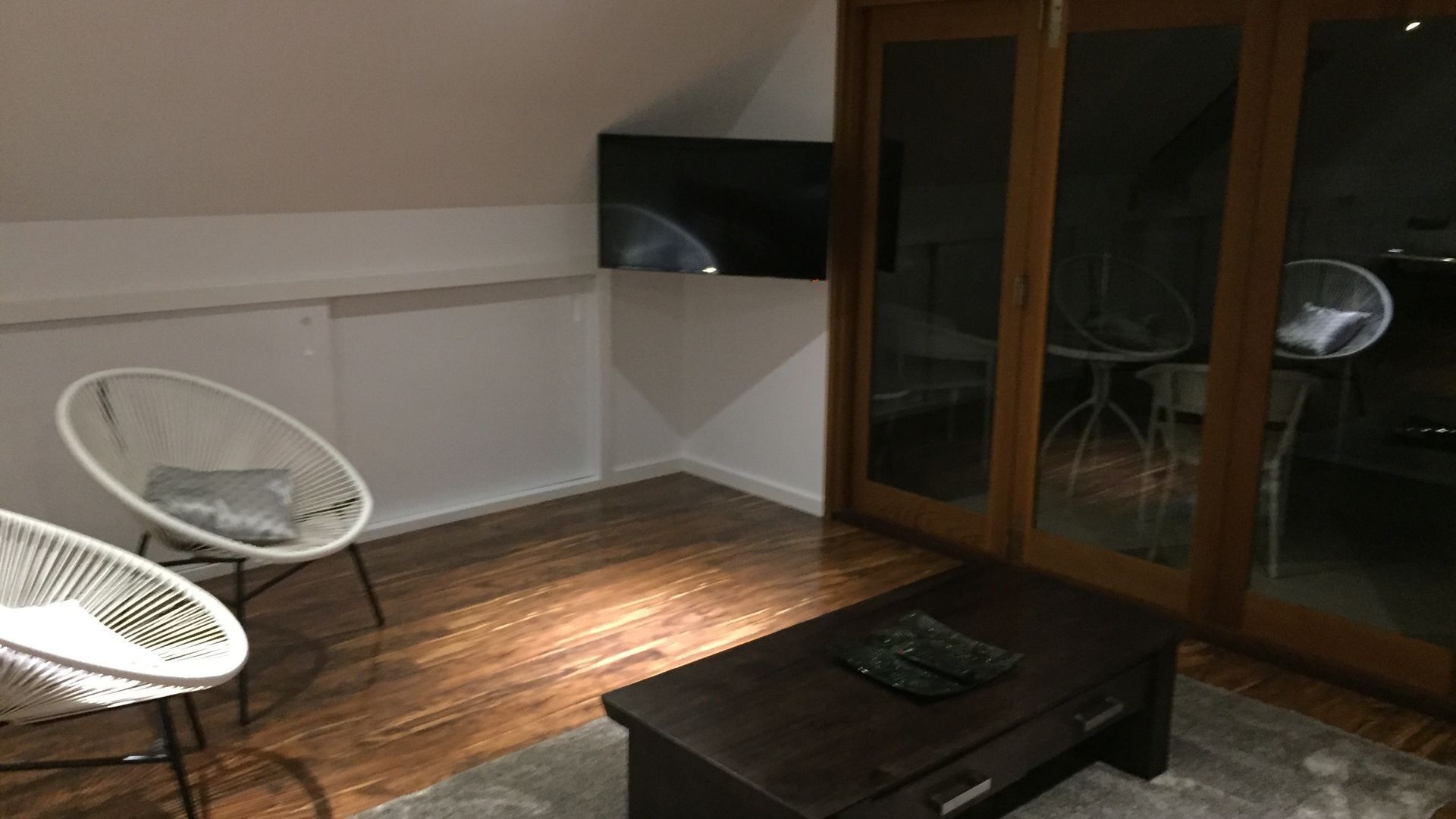 Una habitación con piso de madera oscura, una silla blanca con cojín, una mesita pequeña y un ventanal con vistas al exterior.