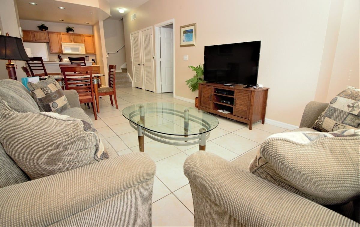 A living room with a glass coffee table, sofas, a television, and a kitchen in the background.