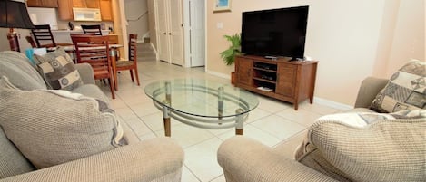 A living room with a glass coffee table, sofas, a television, and a kitchen in the background.