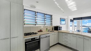 Eine moderne Küche mit Gasherd, Geschirrspüler und Backofen. Ein großes Fenster mit Jalousien bietet Blick auf das Meer.