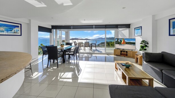 Ein geräumiges Wohnzimmer mit großem Fenster und Blick auf das Meer und die Berge. Es gibt einen Essbereich mit Tisch und Stühlen sowie ein Sofa im Wohnbereich.