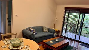 A living room with a sofa, a coffee table, a dining table with a bowl, and a view of greenery through large windows.