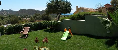 Un patio trasero con césped verde, una silla de madera, un tobogán verde y un árbol.