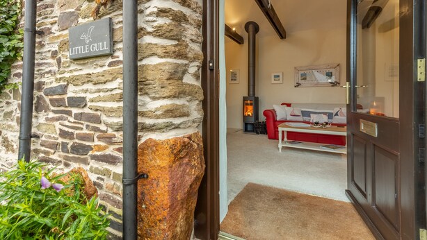 Parede exterior de pedra com um letreiro 'Little Gull', porta de madeira e interior acolhedor com lareira, sofá vermelho e mesa branca.